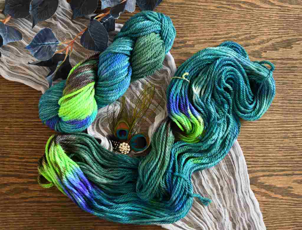 A twisted and an unfolded skein of Barn Yarn hand-dyed bulky weight yarn in colorway Play the Peacock -vibrant teal, emerald green, rich blue inspired by the colors of a peacock’s feathers.