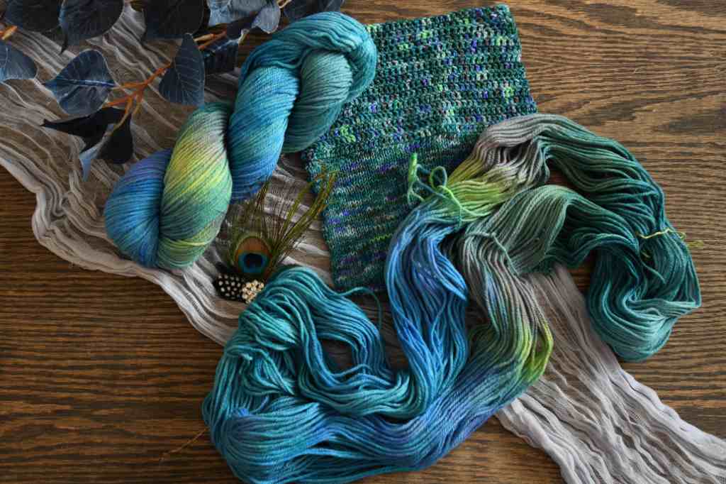 A twisted skein of Barn Yarn hand-dyed worsted weight yarn and an unfolded skein with a sample of knit and crochet fabric in colorway Play the Peacock -vibrant teal, emerald green, rich blue inspired by the colors of a peacock’s feathers.